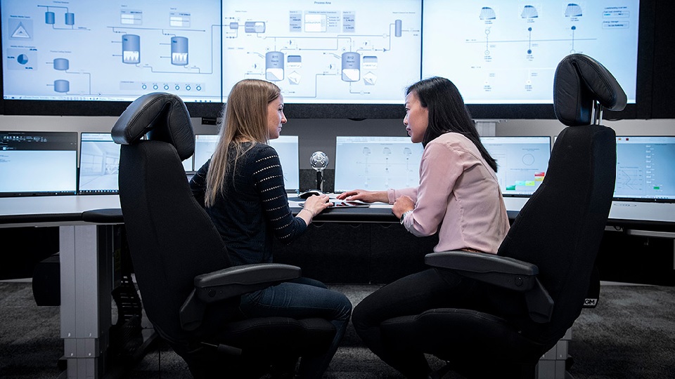Två kvinnor som sitter vid ett skrivbord och pratar. På skrivbordet och på väggen finns flera stora datorskärmar.