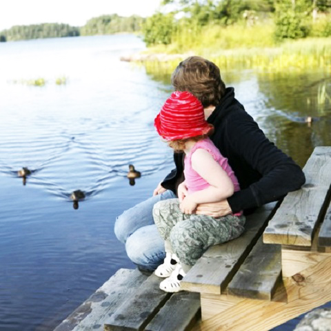 En pappa med ett barn sitter på en brygga och tittar på ankor i vattnet