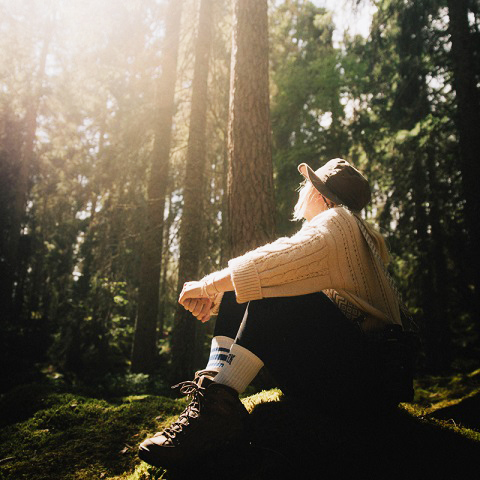 En kvinna sitter på en sten i skogen och tittar upp mot solens strålar som skiner igenom de höga tallarna runt omkring.