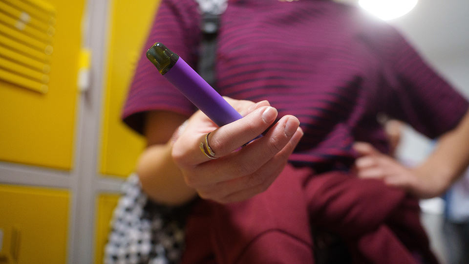 En hand håller en elektronisk cigg. Bakgrunden visar gula skåpluckor.