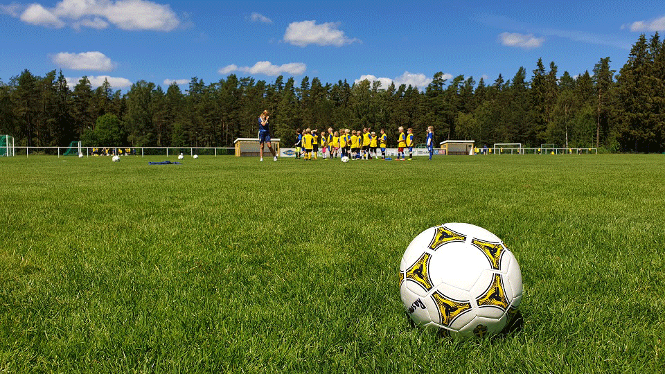 Fotboll på grön gräsmatta med fotbollslag i bakgrunden.