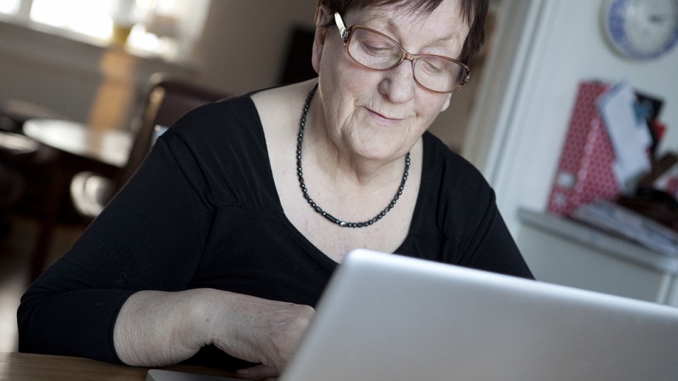 Leende äldre person sitter vid dator och skriver