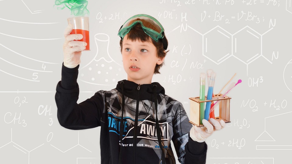 En pojke som i ena handen håller i en behållare med rödfärgad vätska. I andra handen håller han provrör med vätskor i färgerna grön, blå och röd.