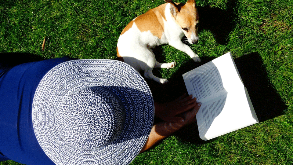 Kvinna med hatt läser bok i gräset tillsammans med hund